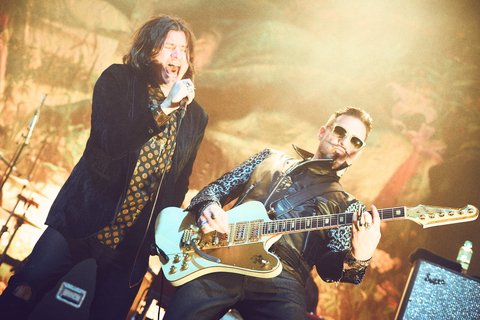 Zwei Sänger treten auf einer Bühne auf. Der linke steht mit Mikrofon und hat lange, dunkle Haare. Der rechte spielt eine elektrische Gitarre und trägt eine Sonnenbrille sowie eine auffällige Jacke. Die Atmosphäre ist lebhaft und energisch.