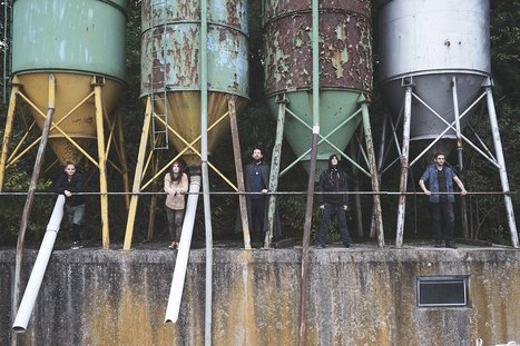 Fünf Personen stehen auf einer Betonfläche vor mehreren großen, farbigen Silos. Die Silos sind rostig und in unterschiedlichen Farbtönen gehalten. Die Menschen wirken entspannt und gehen ihren Aktivitäten nach, während die natürliche Umgebung im Hintergrund sichtbar ist.