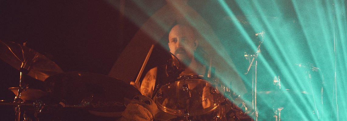 Ein Schlagzeuger spielt energisch auf seiner Drumkit. Im Hintergrund gibt es eindrucksvolle Lichtstrahlen, die eine musikalische Atmosphäre schaffen. Die Szene vermittelt ein Gefühl von Bewegung und Intensität während eines Live-Auftritts.