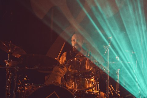 Ein Schlagzeuger spielt energisch auf seiner Drumkit. Im Hintergrund gibt es eindrucksvolle Lichtstrahlen, die eine musikalische Atmosphäre schaffen. Die Szene vermittelt ein Gefühl von Bewegung und Intensität während eines Live-Auftritts.