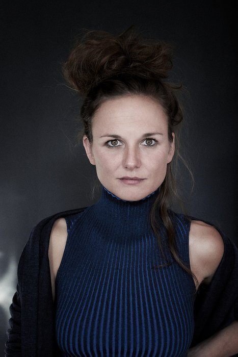 Ein Porträt einer Frau mit einem hochkragen Pullover in blauer Farbe. Sie hat lockiges, hochgestecktes Haar und schaut direkt in die Kamera. Der Hintergrund ist dunkel, was ihren Ausdruck und ihr Outfit betont.