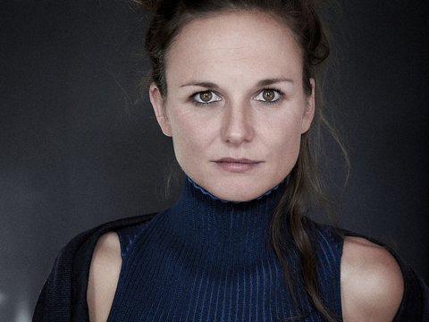 Ein Porträt einer Frau mit einem hochkragen Pullover in blauer Farbe. Sie hat lockiges, hochgestecktes Haar und schaut direkt in die Kamera. Der Hintergrund ist dunkel, was ihren Ausdruck und ihr Outfit betont.