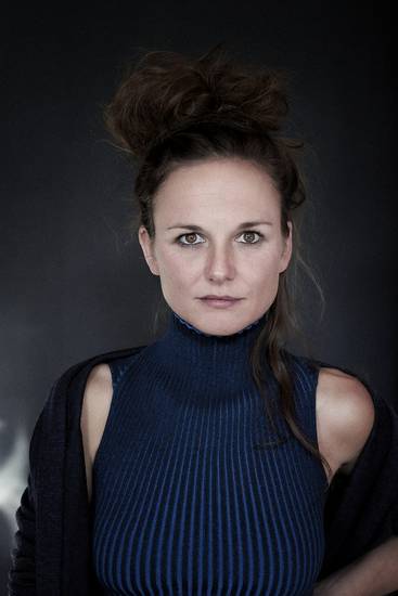 Ein Porträt einer Frau mit einem hochkragen Pullover in blauer Farbe. Sie hat lockiges, hochgestecktes Haar und schaut direkt in die Kamera. Der Hintergrund ist dunkel, was ihren Ausdruck und ihr Outfit betont.