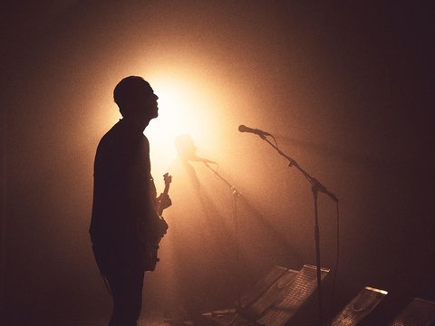 Silhouette eines Musikers im Halbdunkel, der eine Gitarre hält. Im Hintergrund ist eine weitere Silhouette zu sehen. Weiches Licht strahlt hinter ihnen und erzeugt eine stimmungsvolle Atmosphäre. Mikrofonständer sind ebenfalls sichtbar.