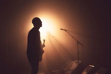 Silhouette eines Musikers im Halbdunkel, der eine Gitarre hält. Im Hintergrund ist eine weitere Silhouette zu sehen. Weiches Licht strahlt hinter ihnen und erzeugt eine stimmungsvolle Atmosphäre. Mikrofonständer sind ebenfalls sichtbar.