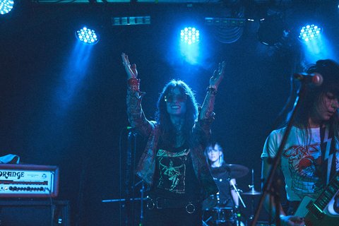 Eine Band spielt auf einer Bühne mit blauen Lichtern. Die Sängerin steht vorn mit erhobenen Händen und lächelt, während die Musiker hinter ihr Instrumente spielen. Die Atmosphäre ist lebhaft und energiegeladen.