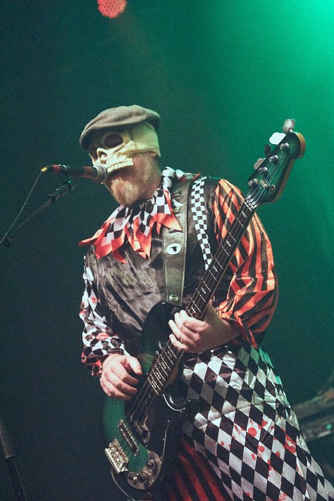 Ein Musiker spieltBass und trägt ein auffälliges Kostüm mit Karomuster und einer Maske, die einen Totenschädel darstellt. Er steht vor einem Mikrofon und ist von grünen Lichtern beleuchtet, während er auf der Bühne performt.