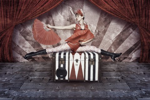 Eine Akrobatin in einem roten Kostüm sitzt auf einem gestreiften Koffer. Sie hält einen Fächer in der Hand und zeigt eine Dehnungsübung. Der Hintergrund ist dekorativ mit roten Vorhängen und gestreiften Elementen gestaltet, was eine zirkusähnliche Atmosphäre schafft.