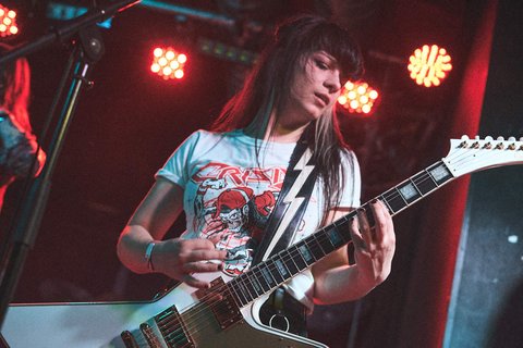 Eine Musikerin spielt leidenschaftlich Gitarre auf der Bühne. Sie trägt ein T-Shirt mit einem auffälligen Design und hat lange, dunkle Haare. Rote und orangefarbene Lichter strahlen um sie herum, während sie sich auf ihren Auftritt konzentriert.