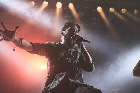 Ein Sänger steht auf der Bühne und hält ein Mikrofon. Er hat eine leidenschaftliche Pose eingenommen und scheint das Publikum mit seiner Performance zu fesseln. Im Hintergrund sind Bühnenlichter zu sehen, die eine dynamische Atmosphäre schaffen.