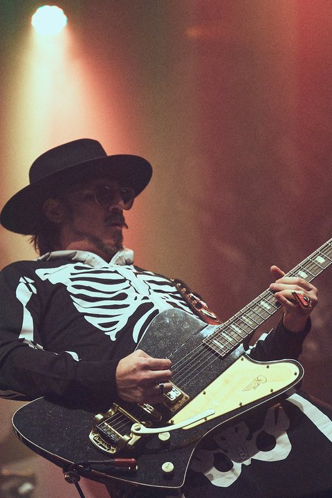 Ein Musiker trägt ein Skelettkostüm und einen schwarzen Hut, während er eine E-Gitarre spielt. Die Bühnenbeleuchtung erzeugt eine stimmungsvolle Atmosphäre.