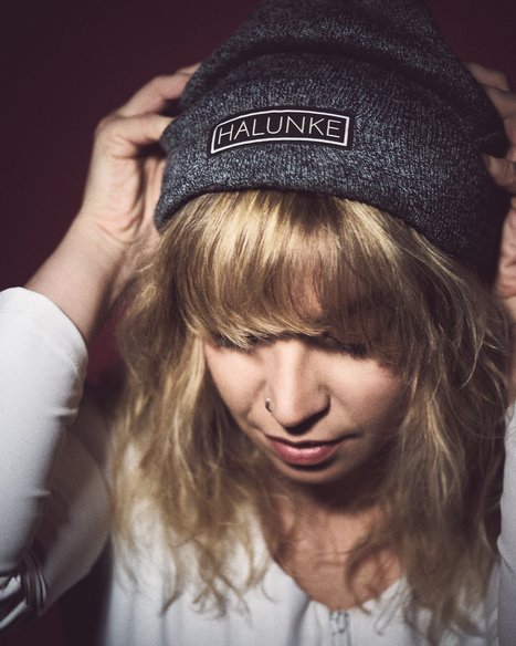 Blonde Woman Adjusting 'HALUNKE' Beanie A young person with wavy blonde hair is wearing a dark beanie and adjusting it. They appear thoughtful, with their gaze directed downward. The background is softly blurred, emphasizing the individual and the beanie's logo.