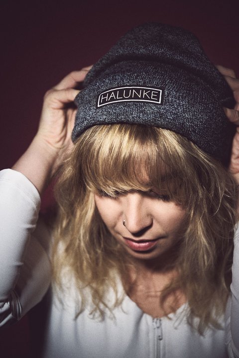 A young person with wavy blonde hair is wearing a dark beanie and adjusting it. They appear thoughtful, with their gaze directed downward. The background is softly blurred, emphasizing the individual and the beanie's logo.