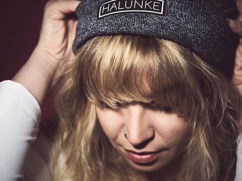 Eine Person mit blonden, lockigen Haaren trägt eine graue Mütze. Sie neigt den Kopf leicht nach unten und berührt die Mütze mit ihren Händen. Der Hintergrund ist dunkel und unscharf, wodurch der Fokus auf der Person liegt.