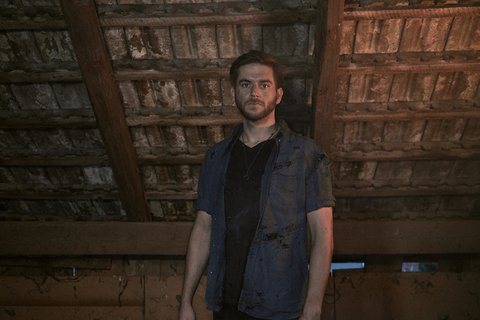 Ein junger Mann steht in einem düsteren, verlassenen Raum mit einer rauen, holzverkleideten Decke. Er hat ernstes Gesicht und trägt ein verwaschenes T-Shirt. Der Boden ist schmutzig, und die Atmosphäre wirkt angespannt und unheimlich.