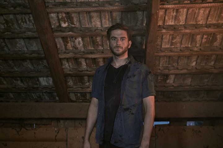 st0nes Ein junger Mann steht in einem düsteren, verlassenen Raum mit einer rauen, holzverkleideten Decke. Er hat ernstes Gesicht und trägt ein verwaschenes T-Shirt. Der Boden ist schmutzig, und die Atmosphäre wirkt angespannt und unheimlich.