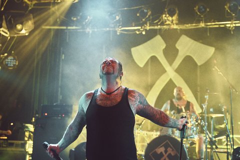 Ein männlicher Sänger mit Tattoos steht auf der Bühne und blickt zum Himmel. Im Hintergrund ist ein Schlagzeuger zu sehen. Die Bühne ist von buntem Licht und Nebel umgeben, während große Äxte als Teil des Bühnenhintergrunds sichtbar sind.