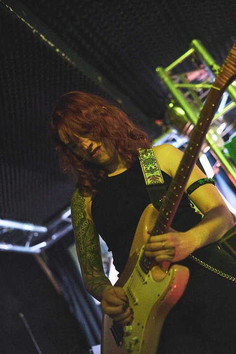 Eine Musikerin mit langen, roten Haaren spielt eine E-Gitarre auf einer Bühne. Sie trägt ein schwarzes Oberteil und hat einen sichtbaren tätowierten Arm. Der Hintergrund ist schwach beleuchtet und zeigt eine Bühnenumgebung.