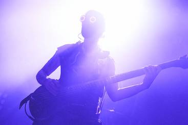 Eine Silhouette einer Person, die einen Bass spielt. Die Person trägt auffällige runde Brillen und steht im Gegenlicht, was eine dramatische Atmosphäre erzeugt. Lila und helle Lichteffekte umgeben die Szene.