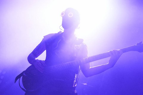 Eine Silhouette einer Person, die einen Bass spielt. Die Person trägt auffällige runde Brillen und steht im Gegenlicht, was eine dramatische Atmosphäre erzeugt. Lila und helle Lichteffekte umgeben die Szene.