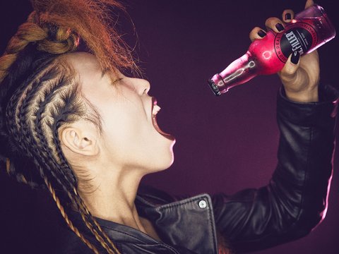 Eine Person mit geflochtenem Haar und schwarzer Lederjacke schreit laut. Sie hält eine pinkfarbene Flasche in der Hand, aus der sie zu trinken scheint. Der Hintergrund ist in einem kräftigen Farbton gehalten und verstärkt die Energie der Szene.
