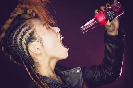 Eine Person mit geflochtenem Haar und schwarzer Lederjacke schreit laut. Sie hält eine pinkfarbene Flasche in der Hand, aus der sie zu trinken scheint. Der Hintergrund ist in einem kräftigen Farbton gehalten und verstärkt die Energie der Szene.
