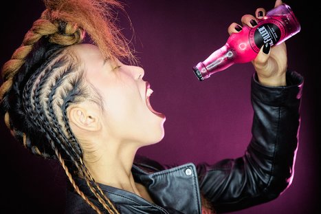 A person with braided hair and a leather jacket is dramatically tilting their head back, mouth open in a shout, while holding a pink soda bottle. The background is dark, highlighting the vibrant colors of the drink and the subject's expressive pose.