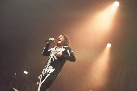 A performer wearing a black skeleton outfit sings passionately on stage. The scene is illuminated by soft, warm stage lights, creating an energetic atmosphere during a live music event. The audience's excitement is palpable in the background.