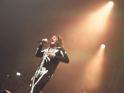 Ein Sänger in einem schwarz-weißen Skelettkostüm steht auf der Bühne und performt. Der Fokus liegt auf seiner energiegeladenen Darbietung, während das Bühnenlicht eine beeindruckende Atmosphäre schafft.