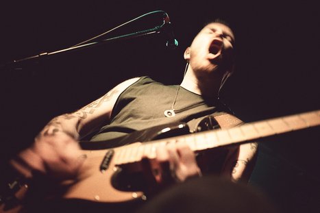 A musician passionately performs on stage, singing into a microphone while playing an electric guitar. They have tattoos visible on their arms and appear focused and energetic, embodying the dynamic atmosphere of a live music event. The background is dark, emphasizing the performer.