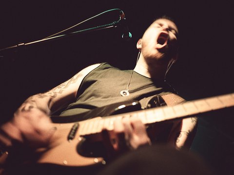 Ein Musiker singt leidenschaftlich in ein Mikrofon, während er eine E-Gitarre spielt. Der Fokus liegt auf seiner energiegeladenen Darbietung und den sichtbaren Tätowierungen auf seinem Arm. Das Bild vermittelt eine intensive, rockige Atmosphäre.