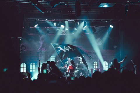 Eine Band steht auf einer Bühne, umgeben von einer begeisterten Menge. Scheinwerfer strahlen hell auf die Musiker, während ein Fan eine Flagge mit einem Logo schwenkt. Die Atmosphäre ist energiegeladen und voller Musikbewegung.