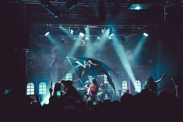 Eine Band steht auf einer Bühne, umgeben von einer begeisterten Menge. Scheinwerfer strahlen hell auf die Musiker, während ein Fan eine Flagge mit einem Logo schwenkt. Die Atmosphäre ist energiegeladen und voller Musikbewegung.