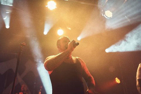 Ein Mann mit Glatze trägt ein schwarzes Tanktop und steht auf einer Bühne. Er hält ein Mikrofon und singt, während Scheinwerferlicht auf ihn strahlt. Die Stimmung ist energetisch und lebhaft, passend zu einem Live-Konzert.