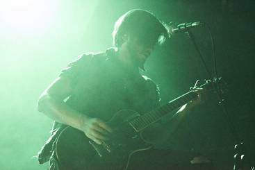 Ein Musiker spielt leidenschaftlich Gitarre auf der Bühne, umgeben von mystischem, grünlichem Licht und Nebel. Seine Aufmerksamkeit ist ganz auf das Instrument gerichtet, während er in die Musik eintaucht.