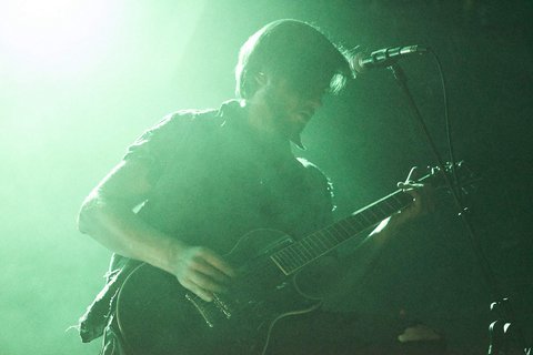 Ein Musiker spielt leidenschaftlich Gitarre auf der Bühne, umgeben von mystischem, grünlichem Licht und Nebel. Seine Aufmerksamkeit ist ganz auf das Instrument gerichtet, während er in die Musik eintaucht.