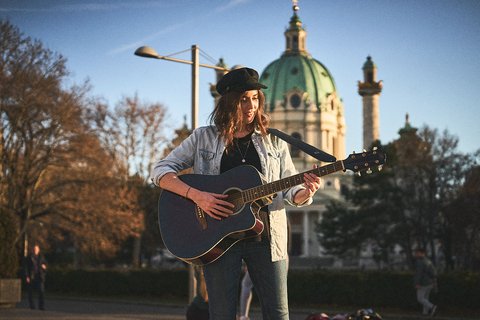 Eine junge Frau spielt Gitarre im Freien. Sie trägt einen Hut und eine Jeansjacke. Im Hintergrund ist eine beeindruckende Kirche mit einer grünen Kuppel und weiteren historischen Gebäuden zu sehen. Es ist ein sonniger Tag.