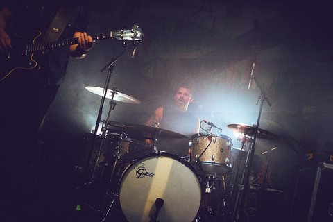 Ein Schlagzeuger spielt in einer schummrigen Umgebung mit aufsteigendem Nebel. Im Vordergrund ist eine Gitarre sichtbar. Die Szene vermittelt eine energiegeladene Atmosphäre eines Live-Konzerts.