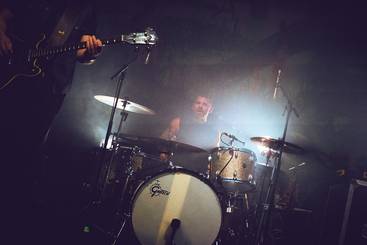 Ein Schlagzeuger spielt in einer schummrigen Umgebung mit aufsteigendem Nebel. Im Vordergrund ist eine Gitarre sichtbar. Die Szene vermittelt eine energiegeladene Atmosphäre eines Live-Konzerts.