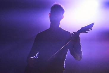 Eine Silhouette eines Mannes, der eine Gitarre hält. Der Hintergrund ist schwach beleuchtet mit violetten Lichtstrahlen. Die Szene vermittelt eine musikalische und dramatische Stimmung.