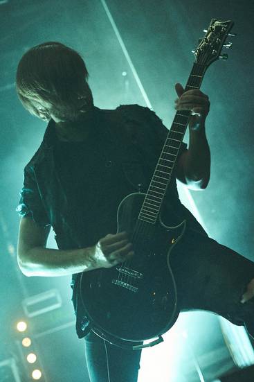 Ein Musiker spielt leidenschaftlich eine E-Gitarre auf einer Bühne. Er trägt ein schwarzes Outfit und hat lange Haare, die teilweise sein Gesicht verdecken. Die Atmosphäre ist dunkel und intensiv mit Beleuchtungseffekten im Hintergrund.
