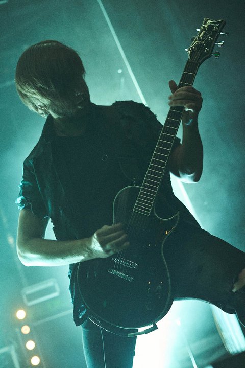 Ein Musiker spielt leidenschaftlich eine E-Gitarre auf einer Bühne. Er trägt ein schwarzes Outfit und hat lange Haare, die teilweise sein Gesicht verdecken. Die Atmosphäre ist dunkel und intensiv mit Beleuchtungseffekten im Hintergrund.