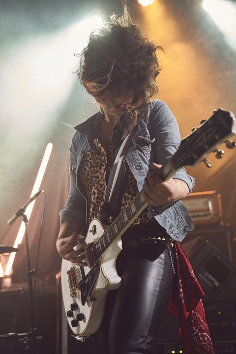 A musician performs passionately on stage, playing an electric guitar with wild energy. They wear a denim jacket over a leopard print shirt and leather pants, with dramatic lighting creating a vibrant atmosphere around them. The focus is on their intense performance and musical expression.