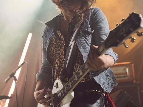 Eine Musikerin spielt Gitarre auf einer Bühne, während Lichter in verschiedenen Farben leuchten. Sie trägt eine Lederjacke und hat einen wilden Haarschnitt. Die Atmosphäre wirkt energiegeladen und lebhaft, passend zu einem Rockkonzert.