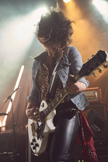 Eine Musikerin spielt Gitarre auf einer Bühne, während Lichter in verschiedenen Farben leuchten. Sie trägt eine Lederjacke und hat einen wilden Haarschnitt. Die Atmosphäre wirkt energiegeladen und lebhaft, passend zu einem Rockkonzert.
