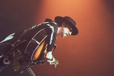 Ein Musiker trägt einen schwarzen Hut und eine auffällige Jacke. Er spielt eine elektrische Gitarre und ist in einer dynamischen Pose. Der Hintergrund ist in warmen Farben beleuchtet, was zur lebendigen Atmosphäre des Konzerts beiträgt.