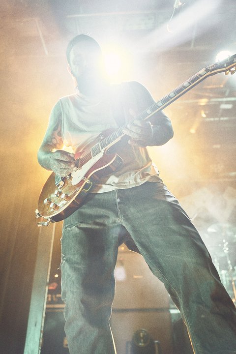 Ein Musiker steht auf der Bühne und spielt leidenschaftlich Gitarre. Ein starkes Licht strahlt hinter ihm und erzeugt eine energiegeladene Atmosphäre. Der Fokus liegt auf dem Musiker und seiner Performance, während der Zuschauerraum unscharf bleibt.