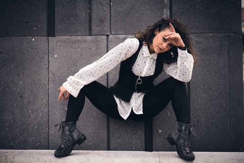 Tessa Eine Person mit lockigen Haaren sitzt in einer tiefen Hocke auf einer Steinoberfläche. Sie trägt ein weißes Hemd mit Punkten und eine schwarze Weste. Ihre Hand ist an der Stirn, als ob sie blinzelnd in die Ferne schaut. Der Hintergrund ist grau und strukturiert.