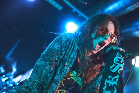 Ein Performer auf einer Bühne singt leidenschaftlich in ein Mikrofon. Er trägt eine auffällige Sonnenbrille und ein buntes, gemustertes Oberteil. Die Hintergrundbeleuchtung erzeugt eine lebendige Atmosphäre, die die Energie des Auftritts unterstreicht.