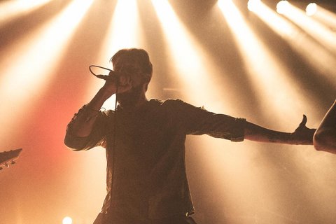 Ein männlicher Sänger steht auf einer Bühne und hält ein Mikrofon, während er leidenschaftlich singt. Sein Arm ist weit ausgestreckt, und er ist von einem warmen Lichtschein umgeben, was die Energie des Live-Auftritts betont. Im Hintergrund ist ein Musiker mit einer Gitarre zu sehen.